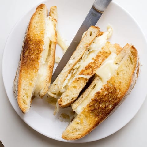 A close-up of a Triple Cheese Sourdough Grilled Cheese sandwich, its golden-brown bread revealing melted cheddar, provolone, and mozzarella on a plate beside a bowl of tomato soup.